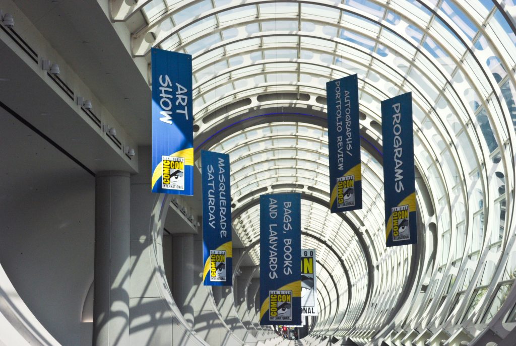 A photo of a bright, modern convention center atrium with a curved glass ceiling and repeating metal arches, lined with tall blue vertical banners advertising San Diego Comic-Con International events such as “Art Show,” “Masquerade Saturday,” “Programs,” “Autographs/Portfolio Review,” and “Bags, Books and Lanyards,” hanging above a long, sunlit corridor.