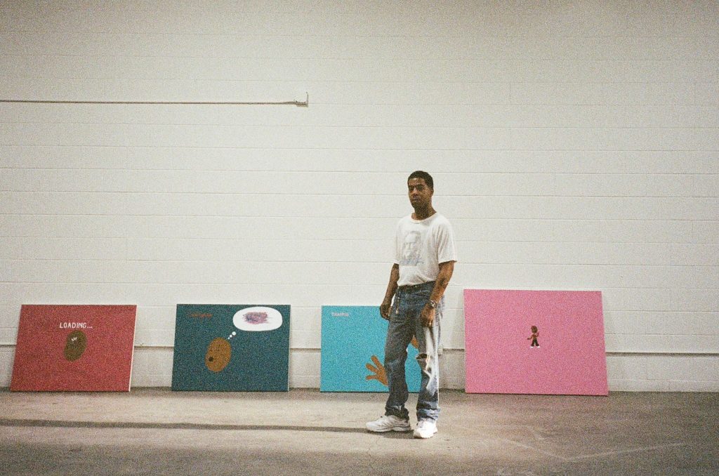 Wide view of a studio, with Scotty Ramon positioned near a row of small, graphic-style paintings arranged along the wal