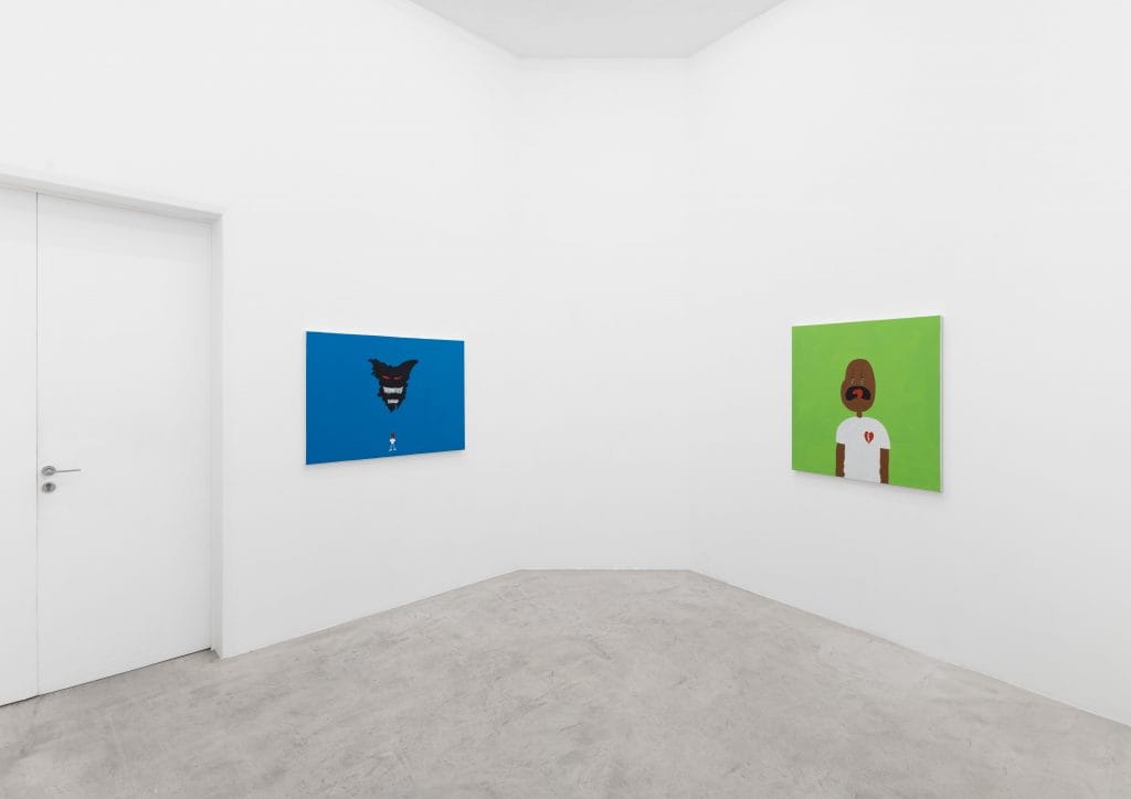 Minimalist white gallery corner with two brightly colored paintings: on the left, a blue canvas featuring a small figure beneath a looming dark creature; on the right, a green canvas showing a stylized figure in a white shirt with a broken-heart emblem.