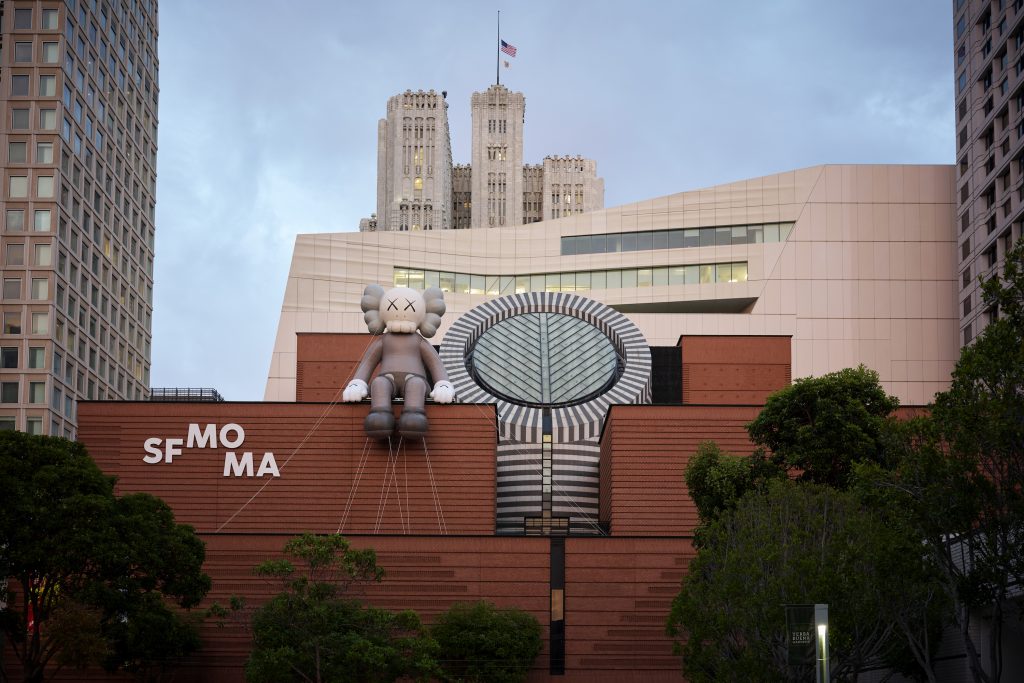 an image of a gray KAWS figure perched on the top of SFMOMA