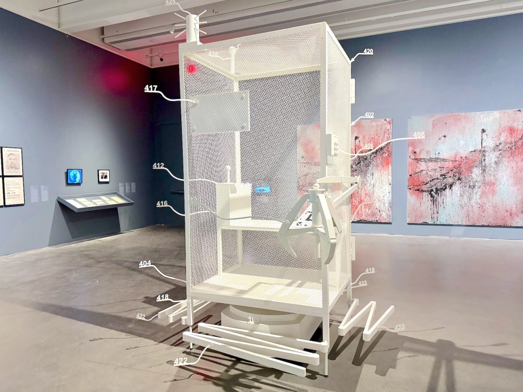 A white mesh cage sculpture with numbered labels and protruding mechanical parts stands in a gallery, positioned in front of two abstract pink and gray paintings.