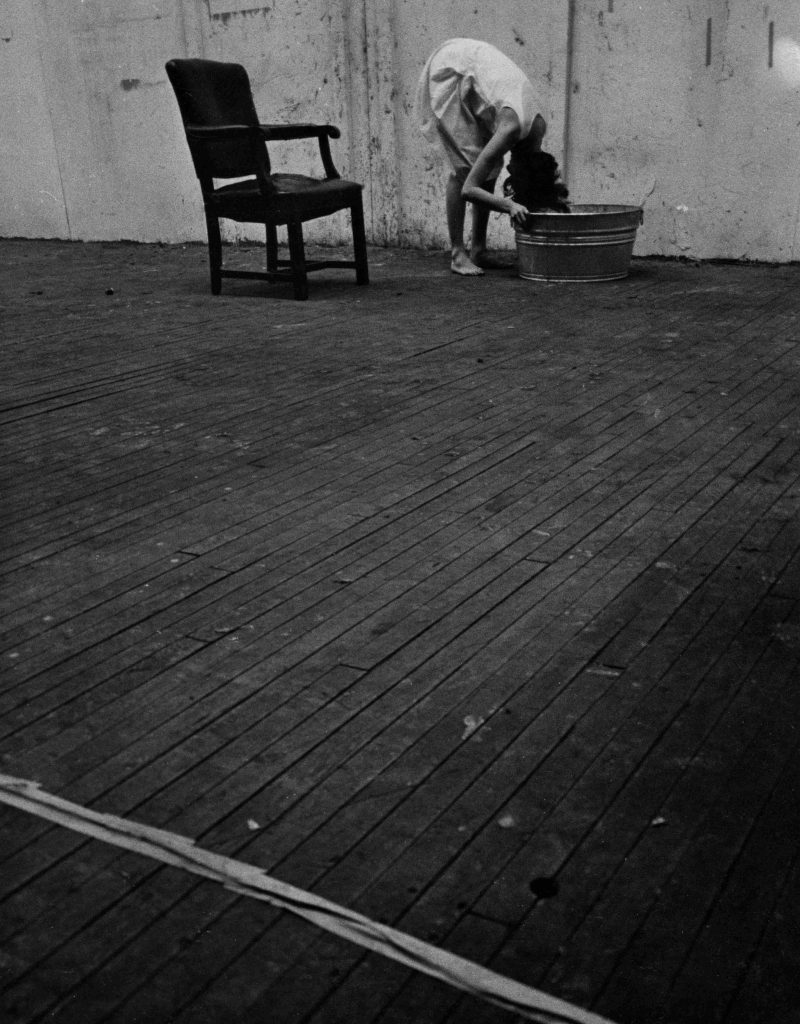 A black and white photograph of a girl with her head in a bucket on an empty wood floor with only a bare-bones old fashioned chair behind her