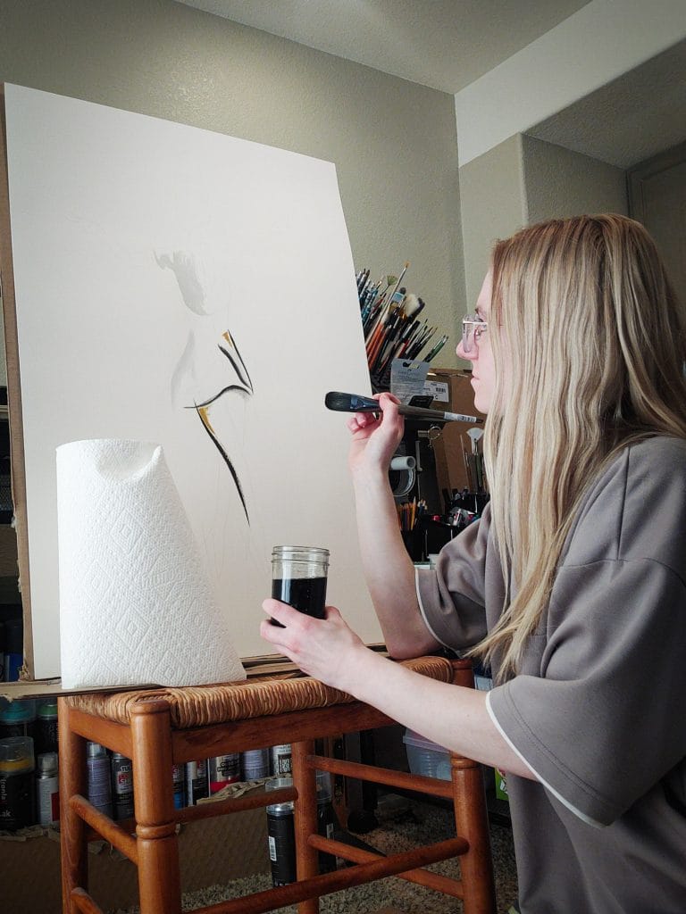 A photo of a young white woman with long blonde hair and glasses sitting at an easel in a home studio, holding a large paintbrush and a jar of dark paint as she works on a minimalist figure painting on a blank white canvas, with art supplies and brushes arranged behind her.
