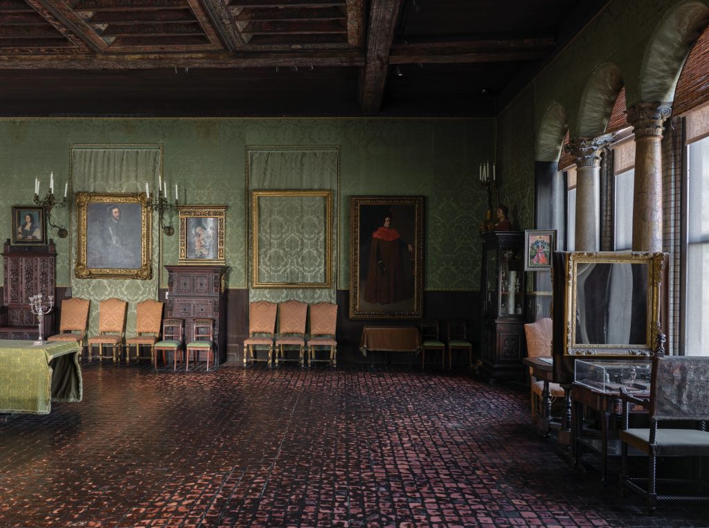 Color photograph of the same historic gallery room, showing green patterned wallpaper, dark wood ceiling beams, and a red-brown tiled floor. Framed paintings, chairs, cabinets, and display cases are arranged along the walls, with arched windows on the right allowing natural light into the space.