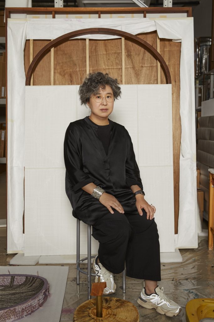 Portrait of an artist seated on a stool in a studio space. The artist has short, curly gray hair, wears round glasses, and is dressed in a loose black outfit with white sneakers. They wear a chunky silver bracelet and a black watch. Behind them is a large wooden frame resembling an arch and wrapped canvas materials. Various sculptural works and studio tools are visible around the workspace.