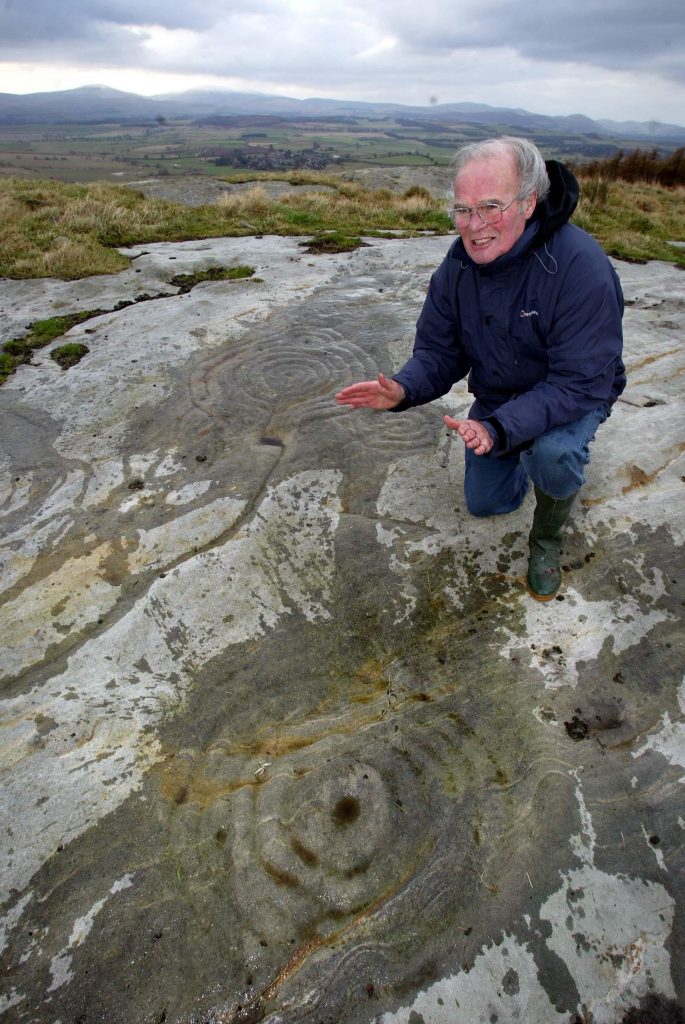 A photo of an older white man with short gray hair and glasses kneeling outdoors on a rocky hillside. He wears a dark jacket, jeans, and green boots and gestures toward large ancient circular carvings etched into the stone beneath him. Rolling fields and distant hills stretch into the background under a cloudy sky.