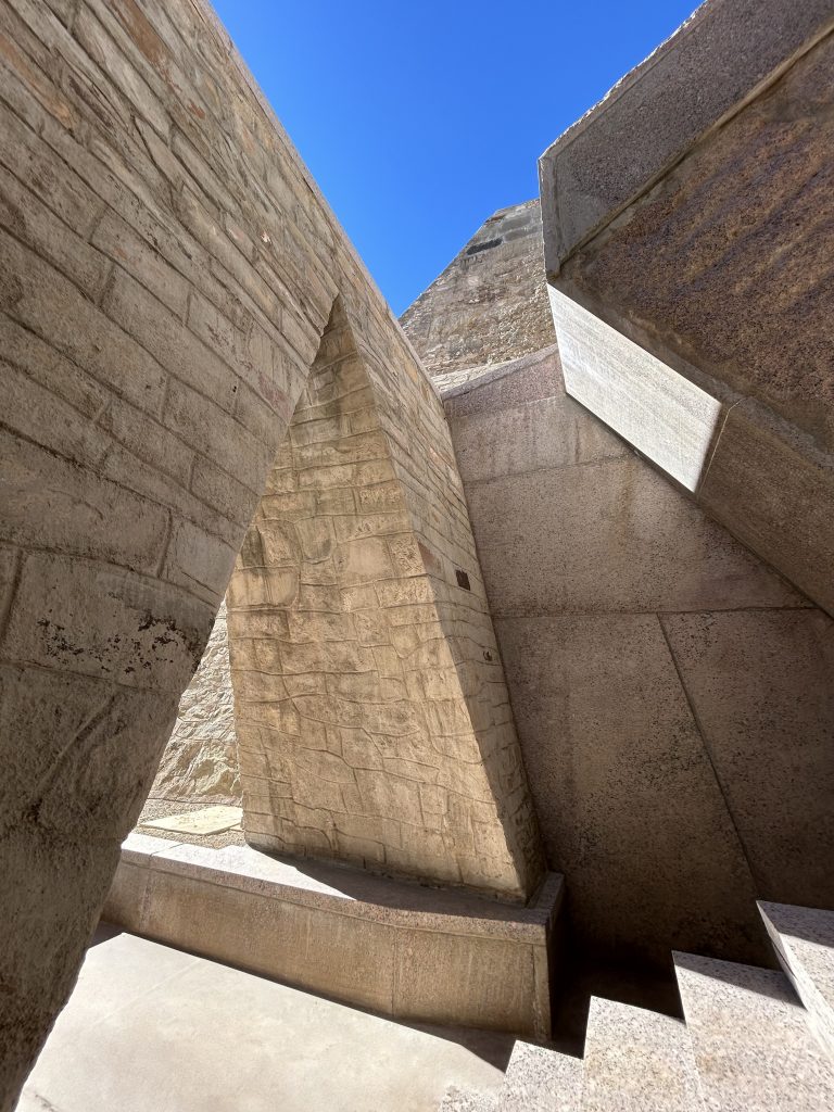 Stone architectural structure with angular walls and stairways under a clear blue sky, viewed from within a geometric passage.