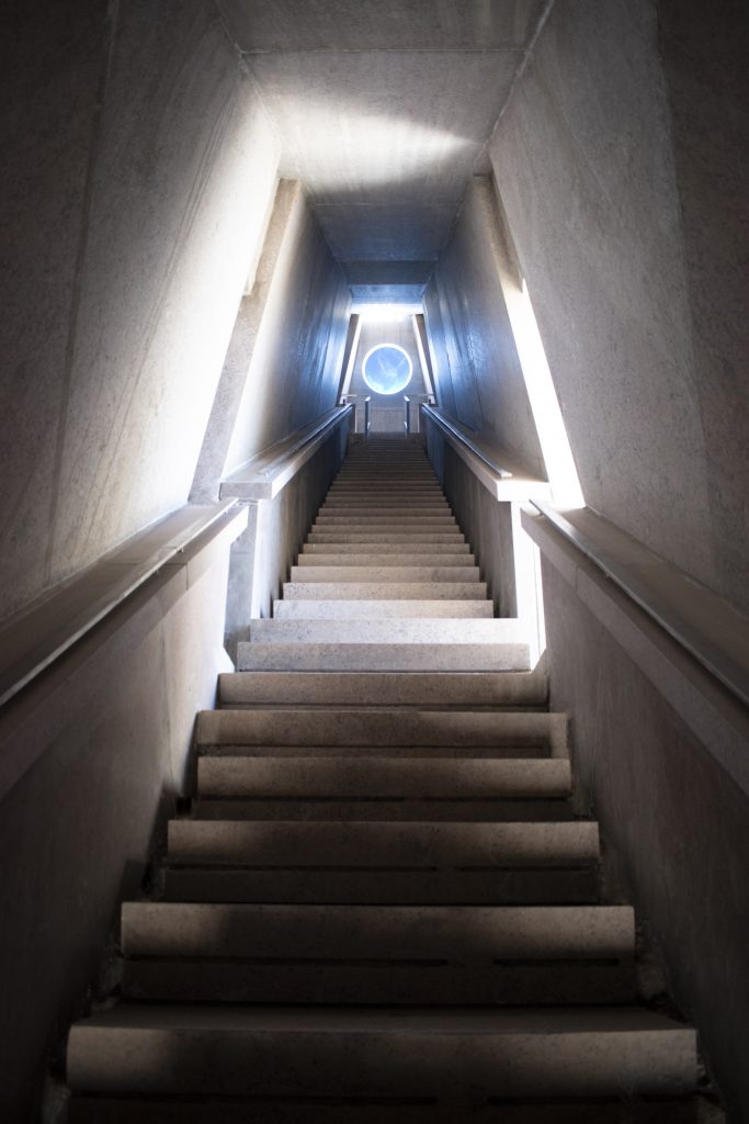 Dramatic stone tunnel with sharply angled triangular walls and central stairway leading toward a circular sky opening.