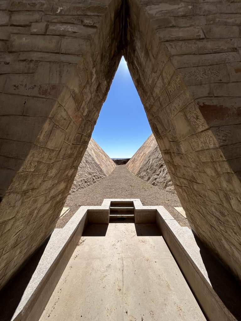 Symmetrical stone corridor with triangular opening framing a long desert vista and bright blue sky.