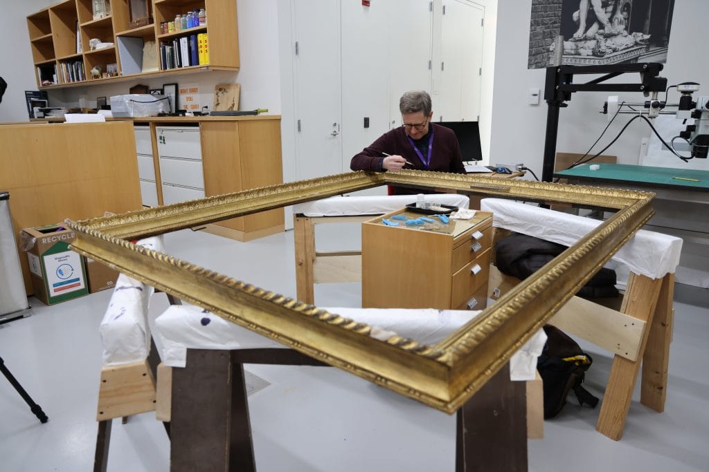 A conservator works on a large, rectangular gilded picture frame resting horizontally on padded supports in a conservation studio. The frame is supported on wooden stands, with tools and materials visible nearby, and worktables and storage cabinets in the background.