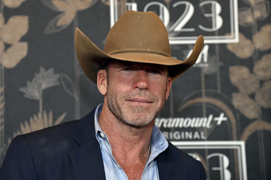 Taylor Sheridan posing in a cowboy hat in front of a backdrop that reads 