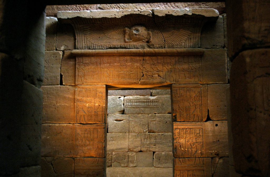 Ancient Egyptian stone wall with carved hieroglyphs and winged sun disk, warmly lit interior temple scene