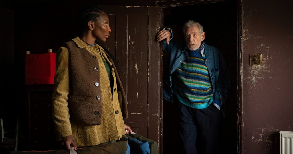 Still of The Christophers, showing two people stand in worn interior doorway, facing each other, one leaning casually, mid-conversation moment