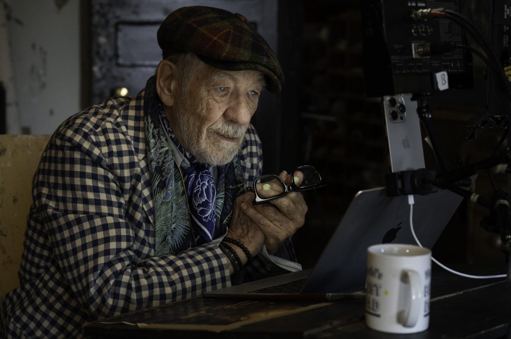 Older man in patterned jacket sits at desk with laptop, holding glasses, surrounded by filming equipment