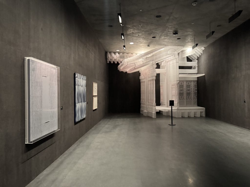 A minimalist museum gallery features textured grey walls, abstract paintings on the left, and a large, translucent fabric sculpture resembling traditional Korean architecture in the background.