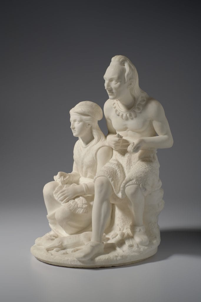 A photo of a white marble sculpture depicting two Native American figures: a middle-aged man with long hair and a necklace seated beside a young girl with braided hair, both in traditional clothing, shown in a calm, intimate pose.