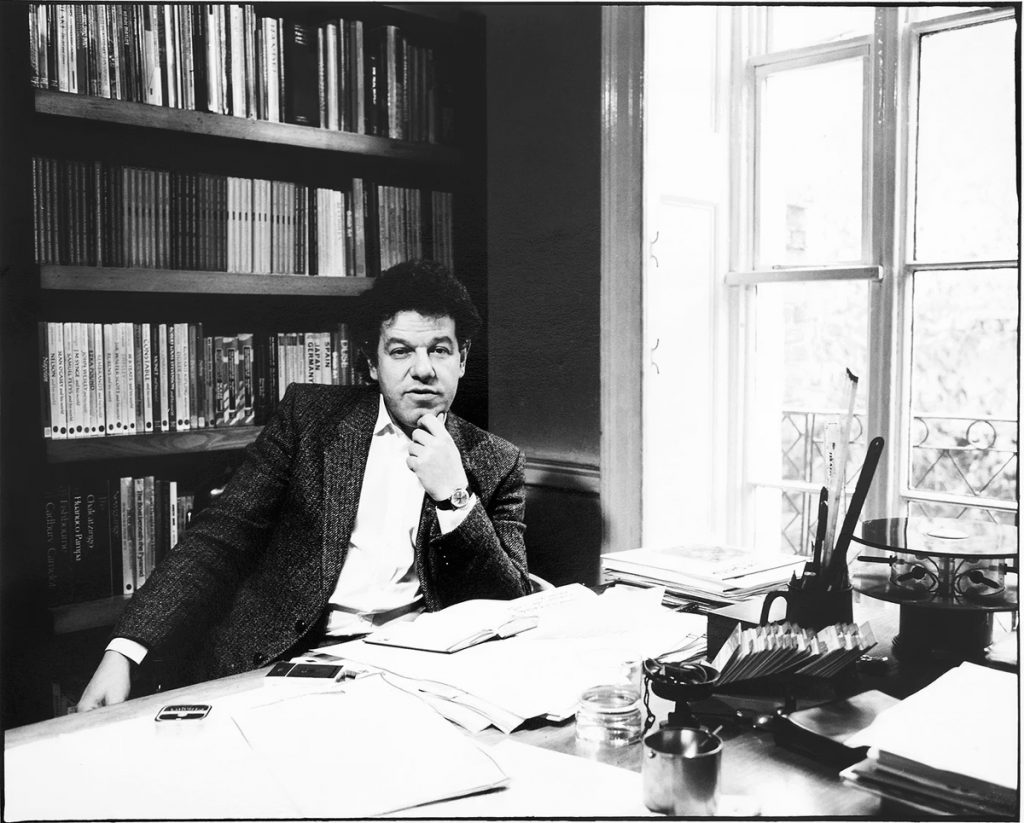 A black and white photo of a young white man with dark curly hair sitting at a cluttered desk in an office. He wears a tweed jacket over a white shirt and rests his chin on his hand while looking toward the camera. Behind him is a tall bookshelf filled with books, and to his side a bright window lets in natural light.