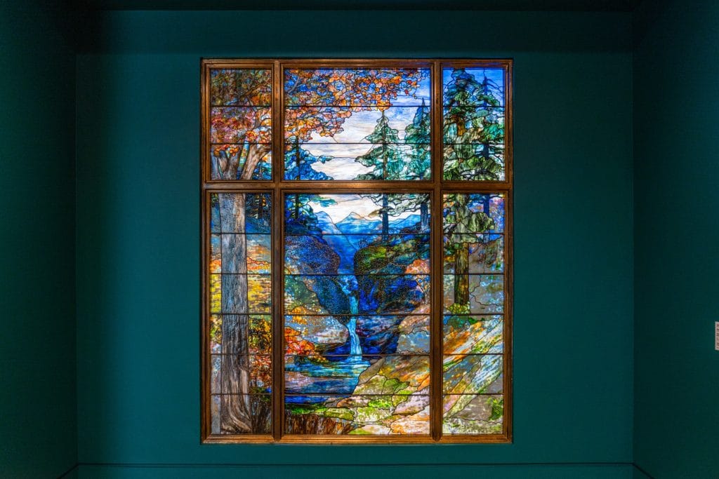 Large Tiffany stained-glass window depicting colorful mountain landscape with trees, waterfall, and vivid sky scenes