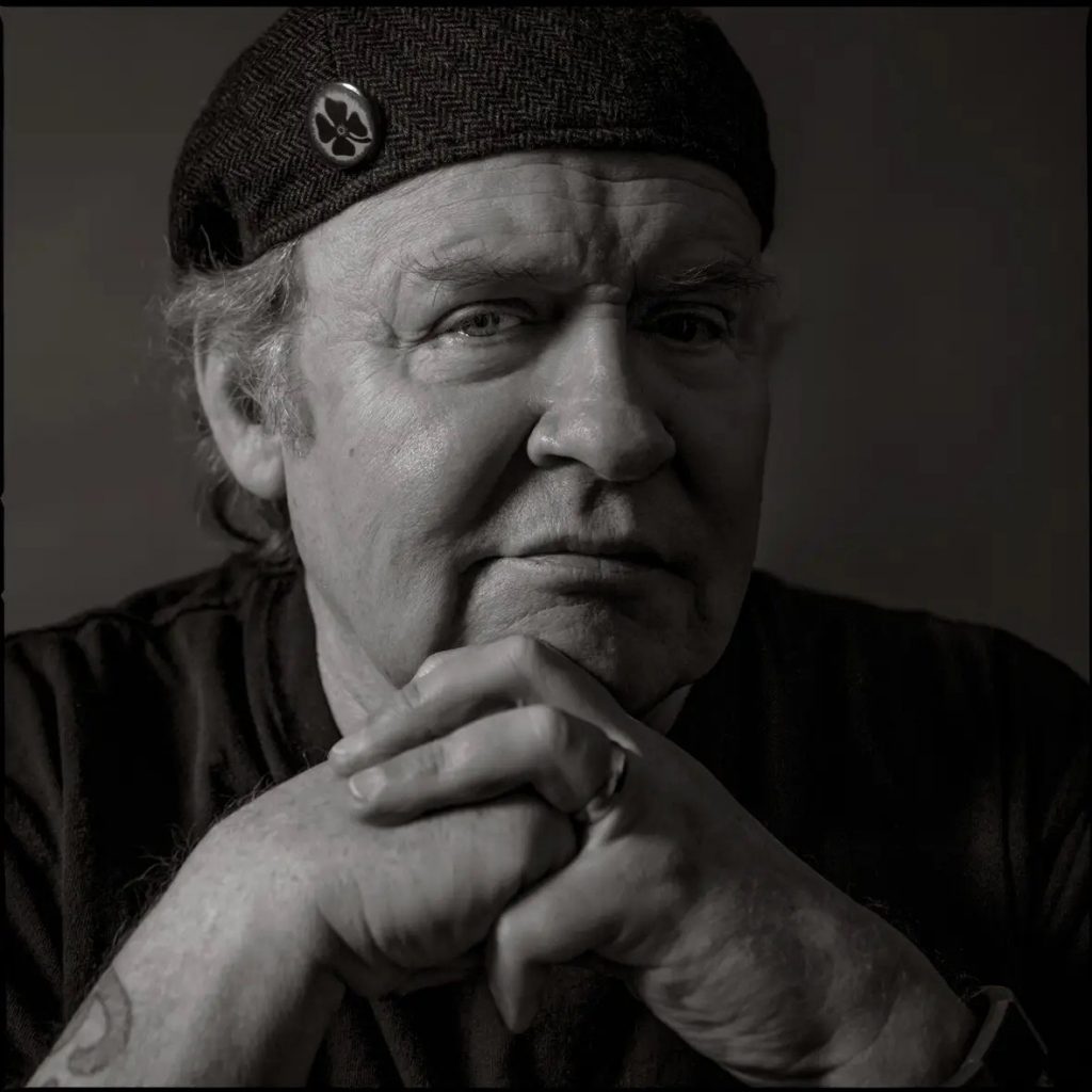 A moodily lit black and photo headshot of a older white man in a black beret, with his hands folded in front of his chin. 