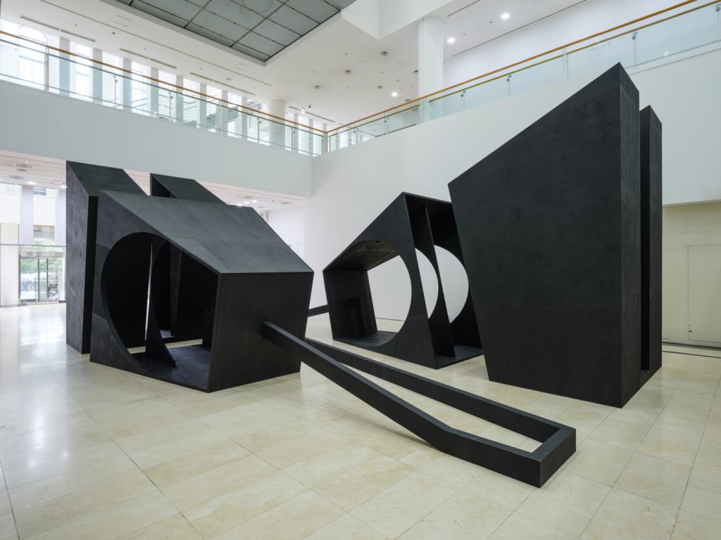 Large-scale, matte black geometric structures with circular cutouts dominate a brightly lit, white-walled gallery space, connected by a long, angular black floor beam.