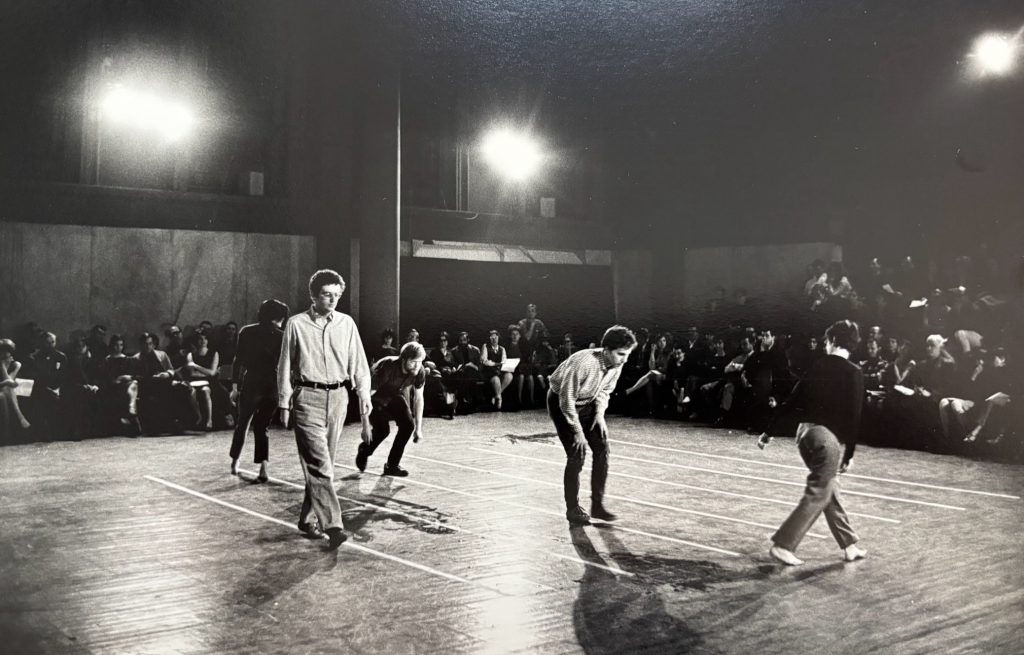 A photograph of five young men playing a game as part of a performance atop lines laid out on the floor, with a crowd surrounding them in chairs and bright stage lights up above it all
