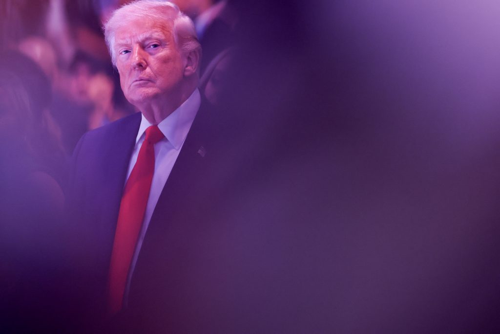 A photo of an older white man with light blond hair and a serious expression, wearing a dark suit and red tie, partially obscured by a purple blur in the foreground at a formal indoor event.