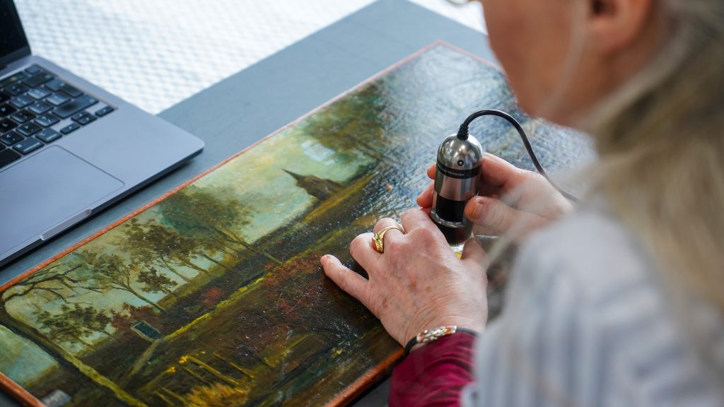 Person uses handheld microscope to examine surface of landscape painting beside laptop on table