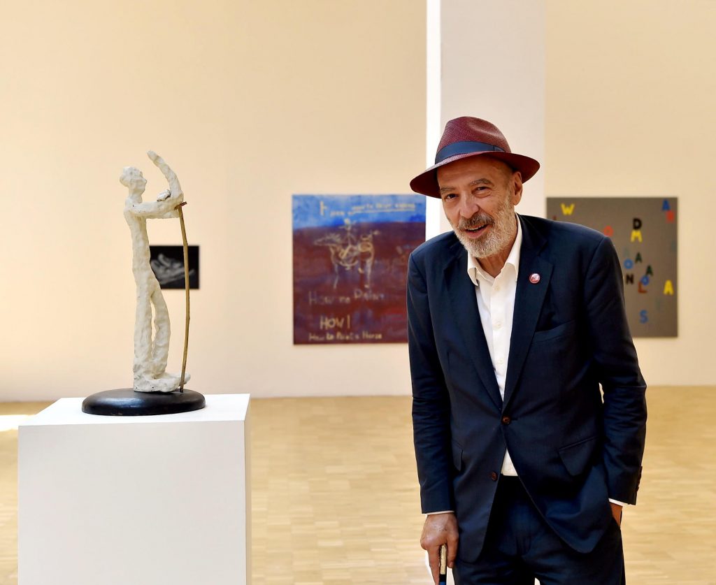 Man in suit and hat stands beside abstract sculpture in gallery with colorful artworks behind.