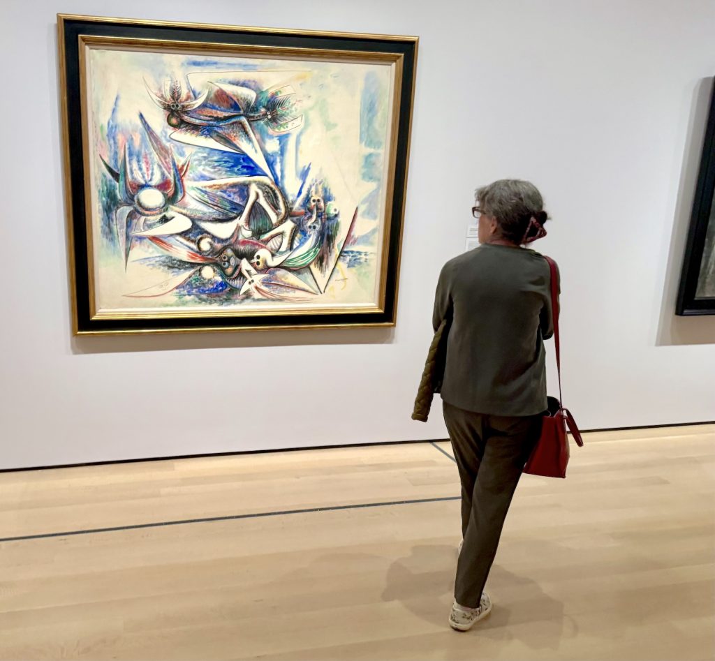 A museum visitor with grey hair and a red shoulder bag stands in a gallery, viewing a large, vibrant painting titled "Chant of Osmosis" by Wifredo Lam. The artwork features a dynamic explosion of cool blues, greens, and sharp, angular forms that suggest bird-like or totemic figures in motion against a pale background.