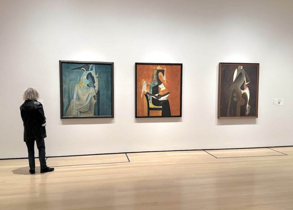 A museum gallery view showing a person standing before three large, surrealist paintings by Wifredo Lam, featuring abstracted, totemic figures against solid-colored backgrounds.