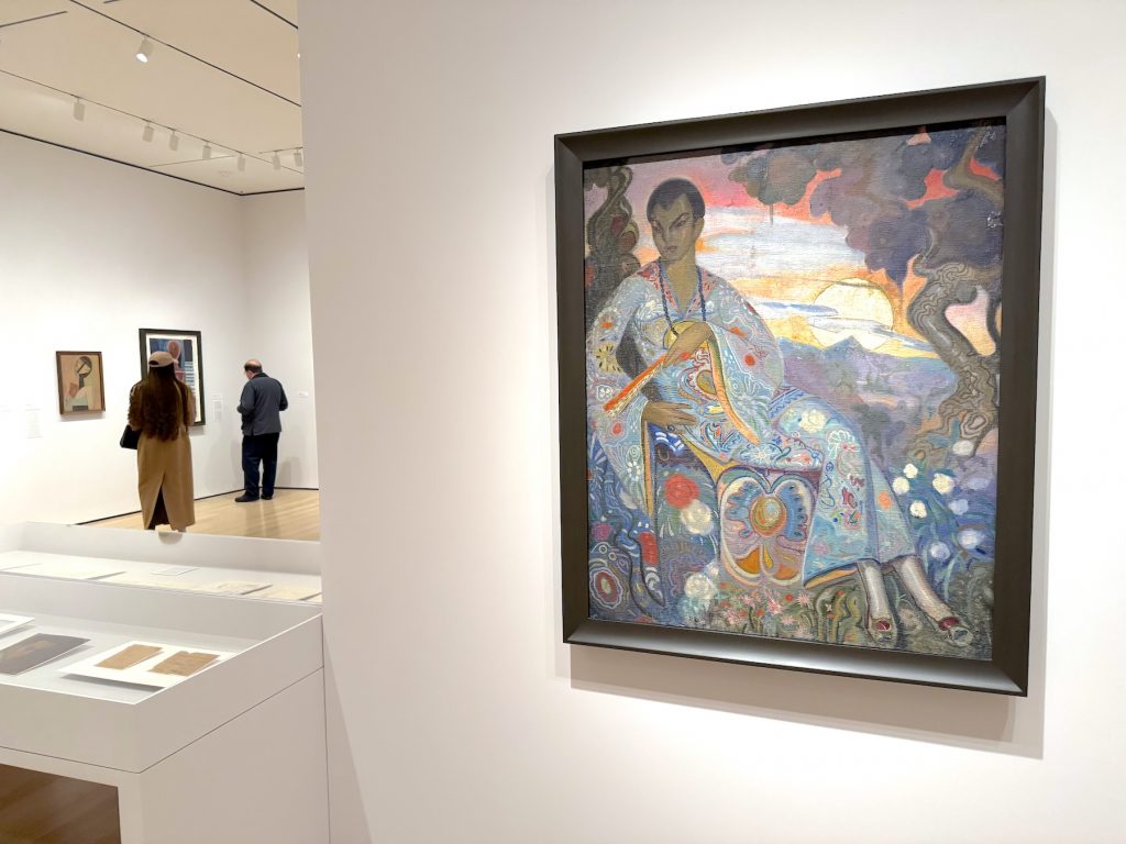 A framed painting by Wifredo Lam displayed in a white-walled gallery, depicting a figure in ornate patterned clothing against a colorful sunset sky with decorative motifs, while two visitors view other artworks in the background.