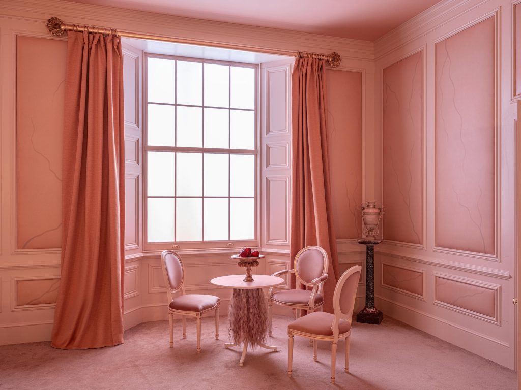 Small pink sitting room with round table, three chairs, tall window, curtains, and decorative molding, from the Airbnb Wuthering Heights experience.