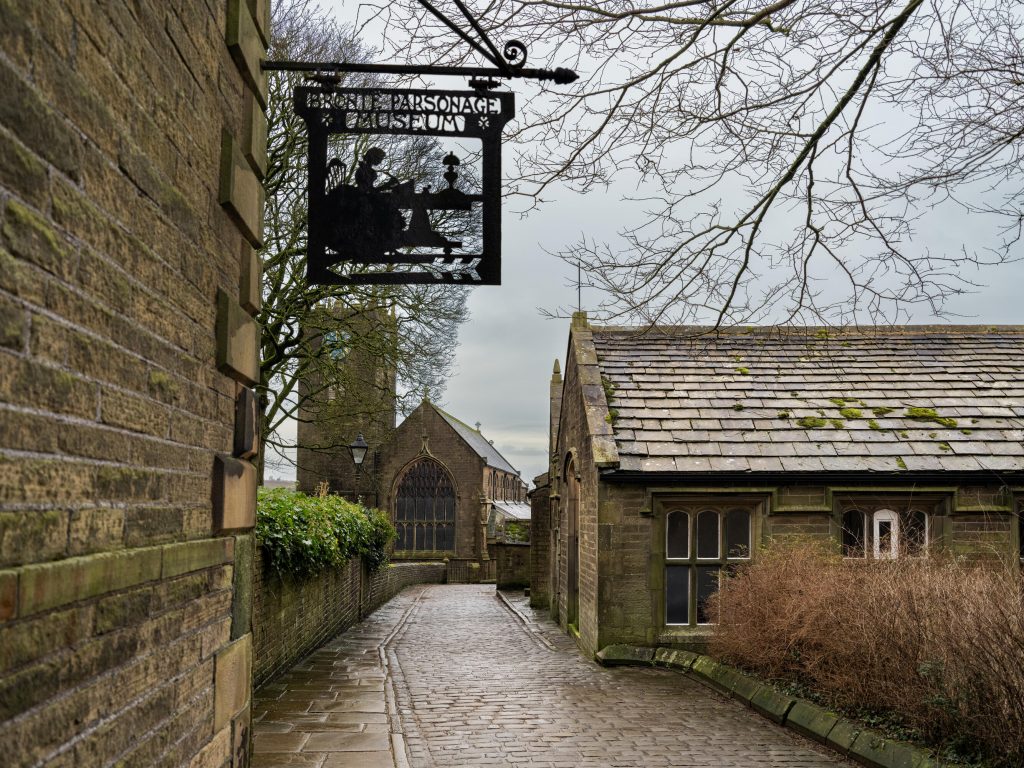 Cobbled lane leads to Brontë Parsonage Museum, stone cottages and church beneath bare trees nearby.
