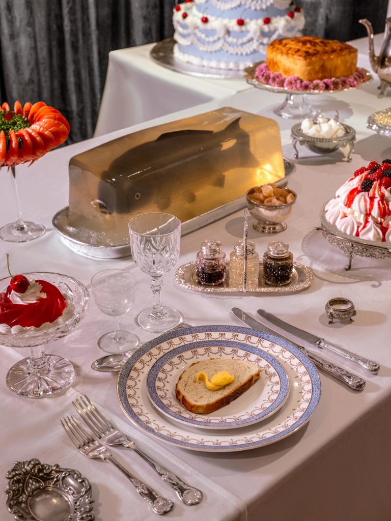 Victorian-style dining table displays fish in aspic, cakes, silverware, and formal place settings arranged meticulously.