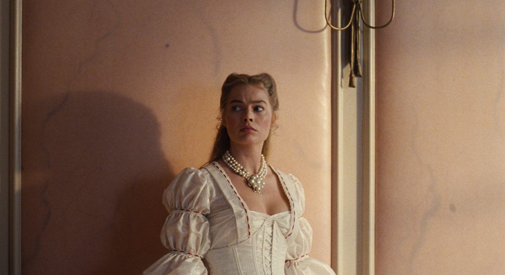 Still from Wuthering Heights showing Margot Robbie in period corset dress stands against pink wall, pearl necklace, gazing tensely off-screen nearby.