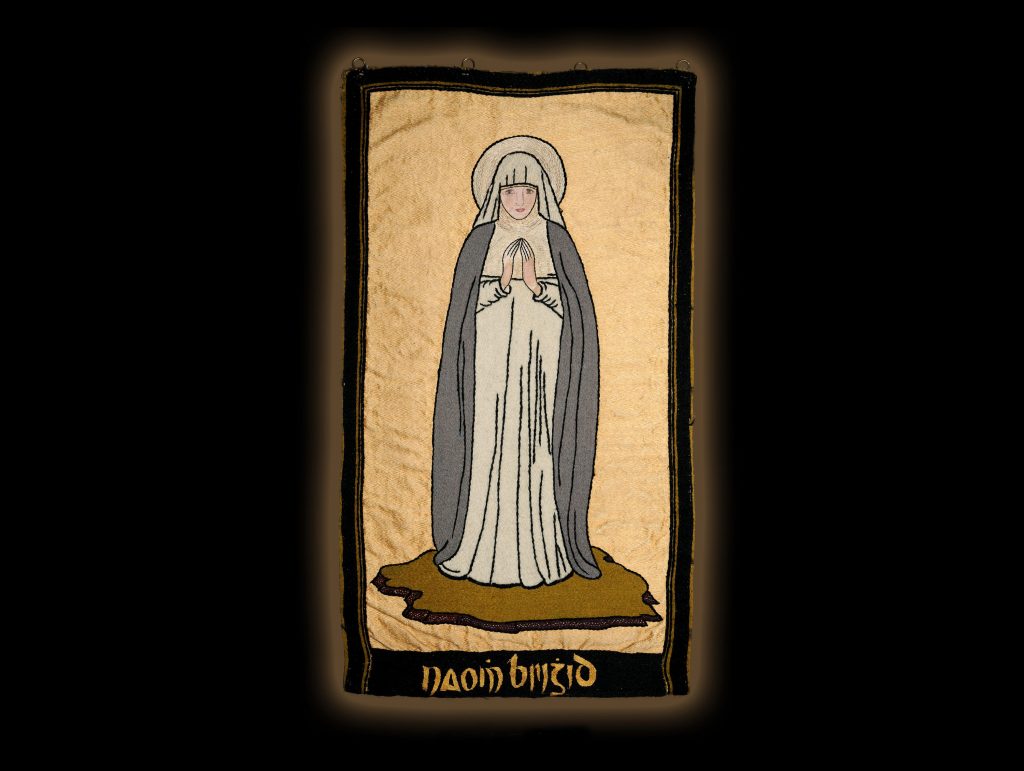 Textile banner depicting Saint Brigid as a haloed nun in white robes and gray cloak, standing with hands clasped in prayer against a plain gold background.