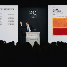 An auctioneer stands at a podium with an abstract painting behind him