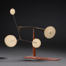 A photograph of the Calder mobile going to auction. A short, red metal 't' shape extends from a red metal base, one yellow disk on one end of its arms and another thinner, bent arm extending off the other, featuring three more discs stationed at each point. All pictured on a gray background