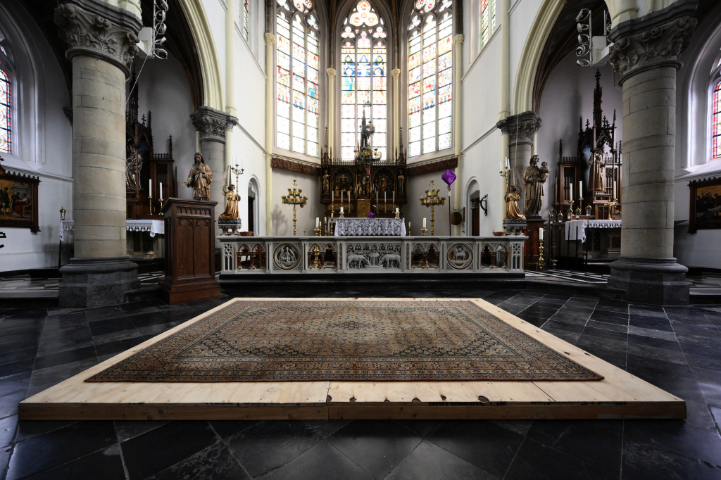 A photograph of a wide open church knave with a triptych of stained glass windows glowing behind the altar—and a wood board with a rug atop it covering the suspected burial of D'Artagnan