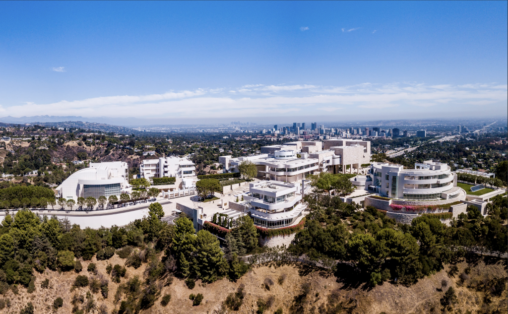 An aerial view of a sprawling white museum campus situated amidst the striking landscape of L.A. on a sunny afternoon
