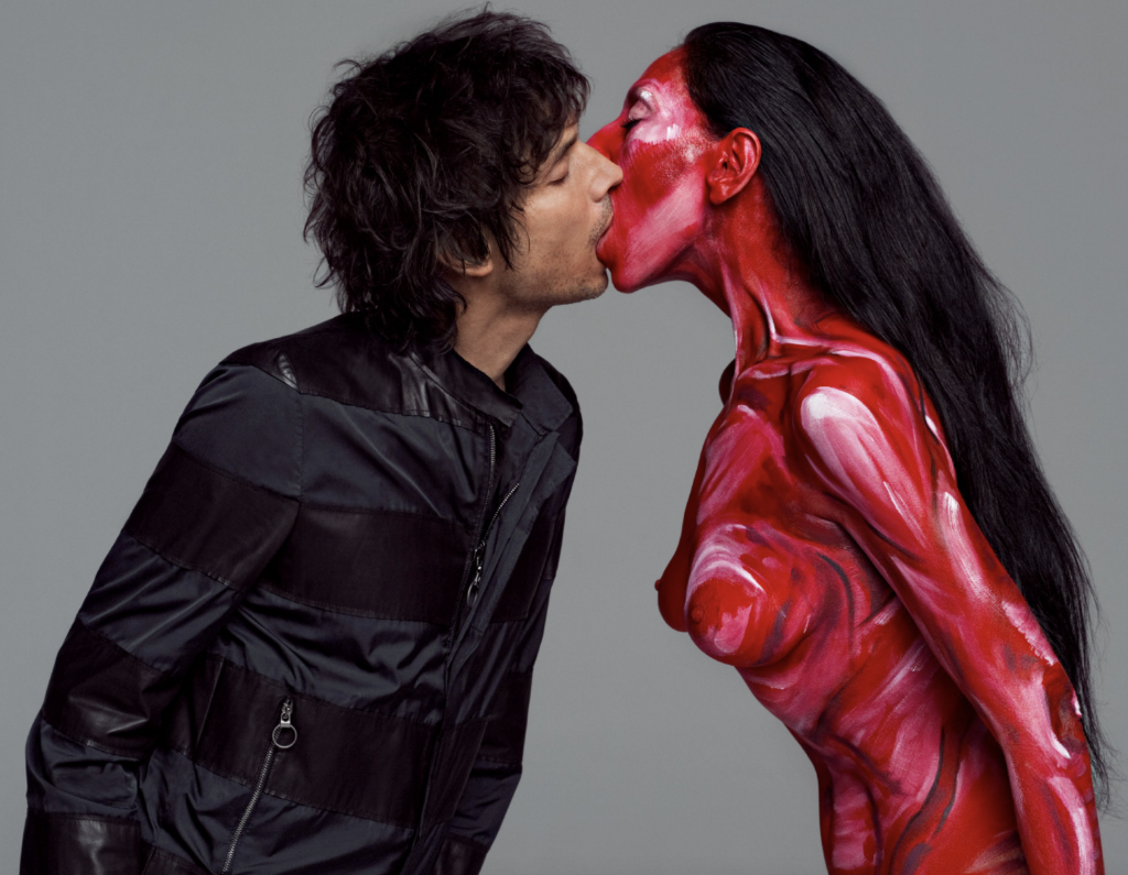 A photograph of Vinoodh wearing a black jacket kissing Inez, covred in red body paint, against a flat gray background