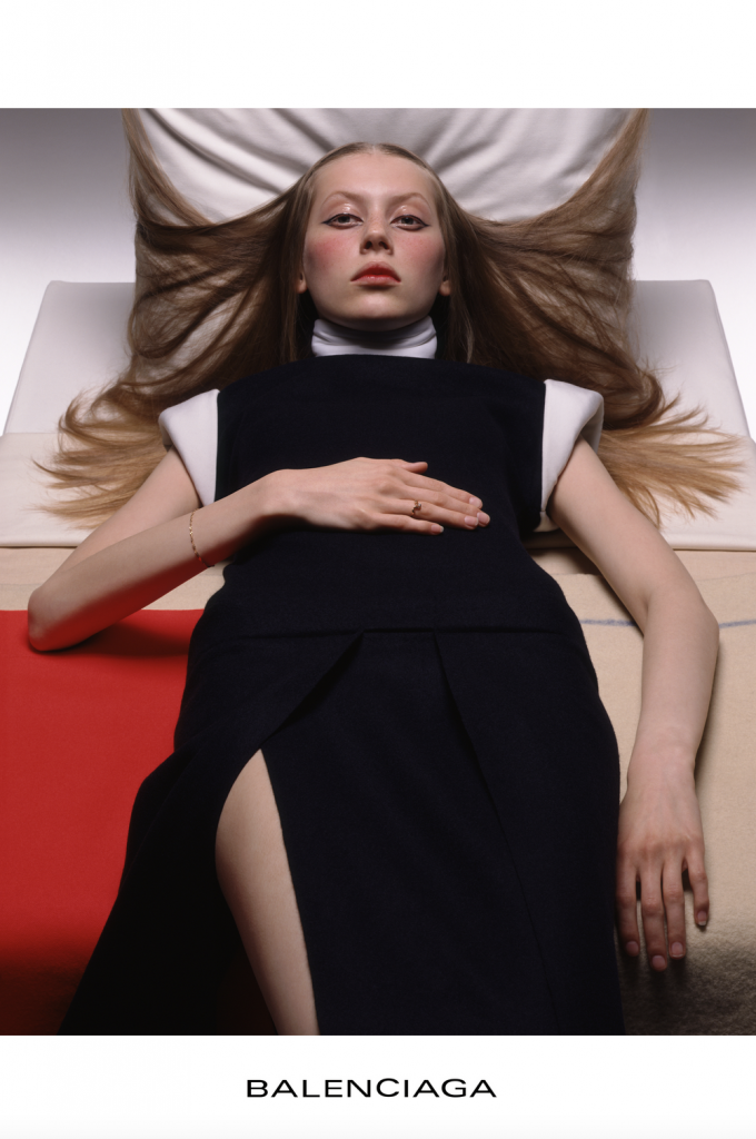 A photograph of a Balenciaga advertisement featuring a young girl in a black dress with white collar and cap sleeves laying on a red and white bed, her blond hair splayed out on the white pillow propped up at an odd angle behind her