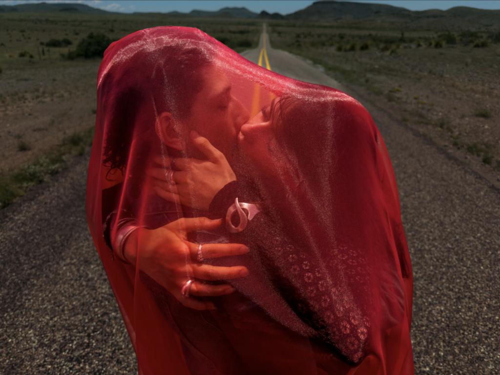 A photograph of two young lovers embracing and kissing beneath a sheer red veil on a long road that stretches out behind them, up and over a hill off in the distance