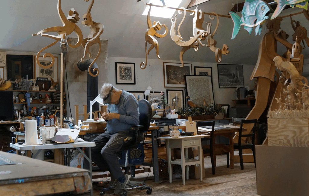 A photo of an older white man with gray hair working intently at a cluttered workbench in a spacious artist’s studio. He wears a magnifying visor and casual clothes while surrounded by tools, papers, and small materials. Overhead hang whimsical wooden sculptures of monkeys and brightly painted fish, and large wooden figures and framed artworks fill the room behind him.