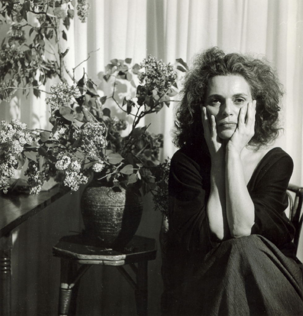 A sepia-toned photograph of Pat Steir, who recently died, seen in her younger years sitting in a chair with hands around her chin next to a big vase of flowers before a white curtain