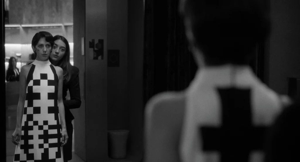 Still from Two People Exchanging Saliva showing two women standing before a mirror in a stark, black-and-white interior; one wears a sleeveless dress with a bold geometric cross pattern while the other stands closely behind her.